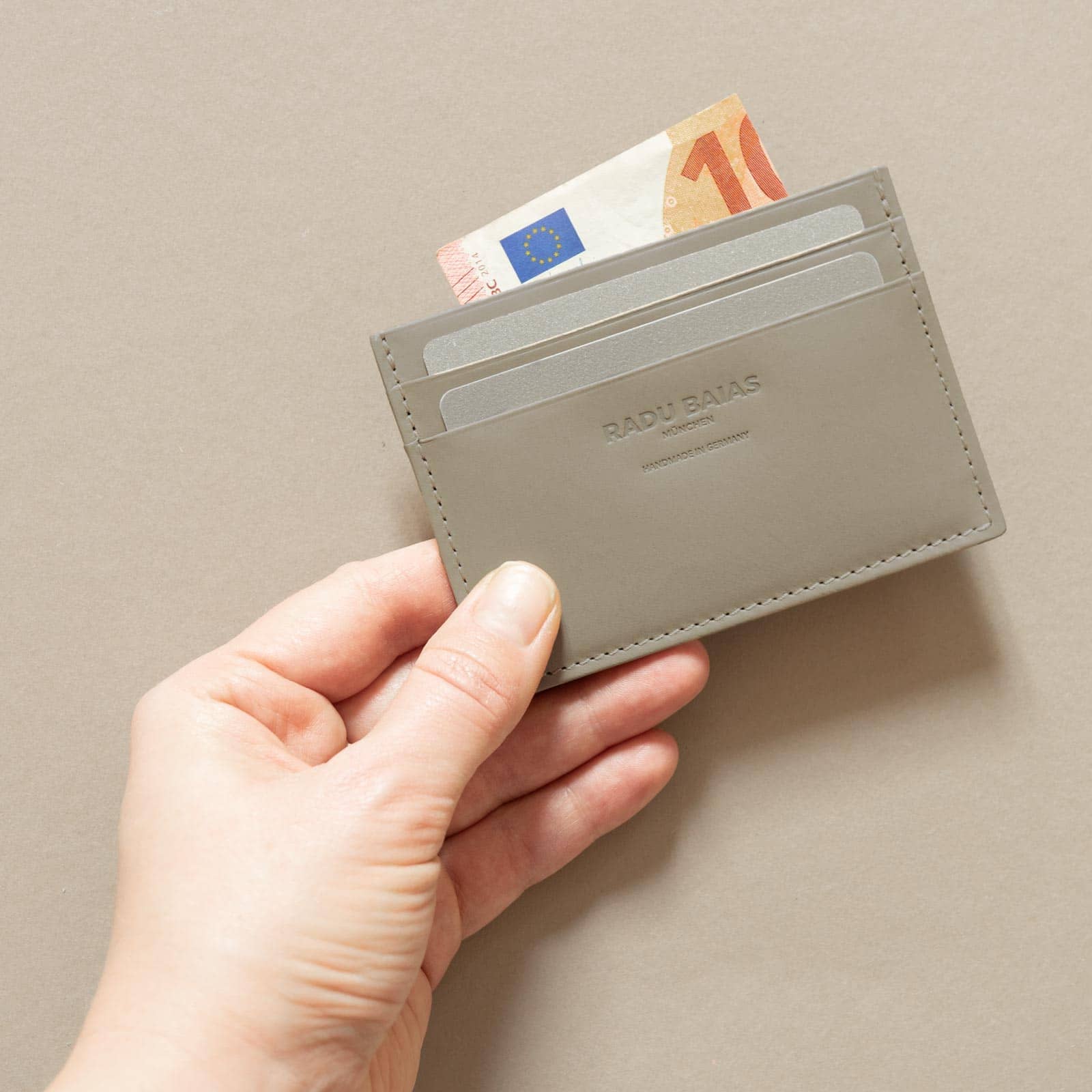 Hand hält Cardholder aus Leder in Moosgrau mit Logoprägung und beispielhaft mit Karten und Geldschein befüllt